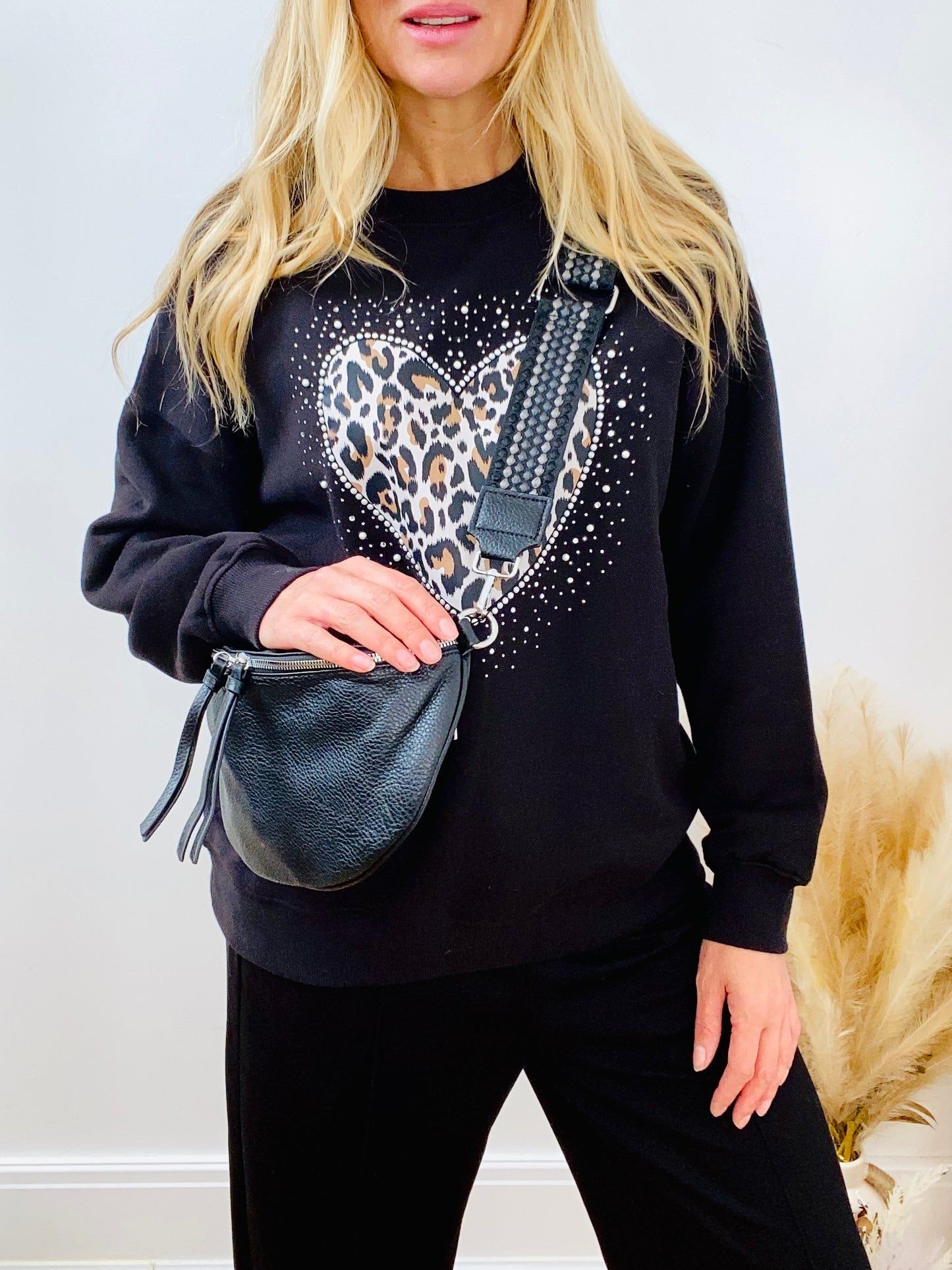 Person wearing a black sweatshirt with a heart design, holding a faux leather black crossbody bum bag