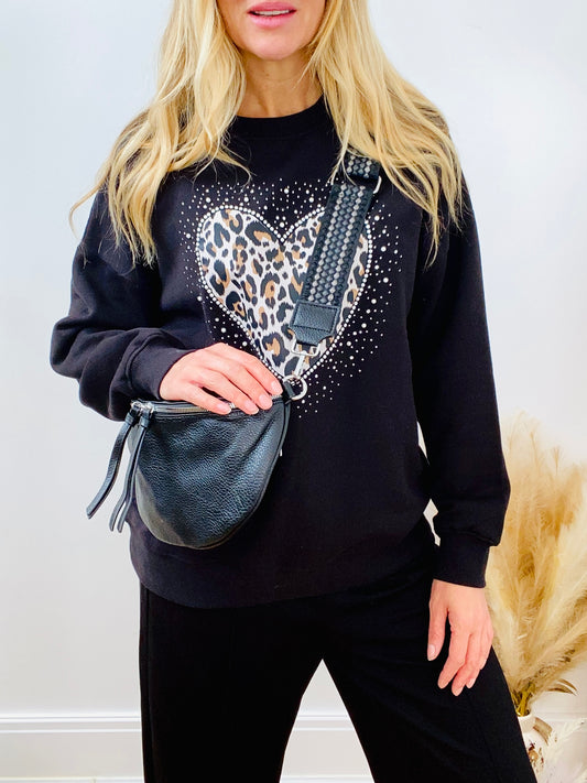 Person wearing a black sweatshirt with a heart design, holding a faux leather black crossbody bum bag