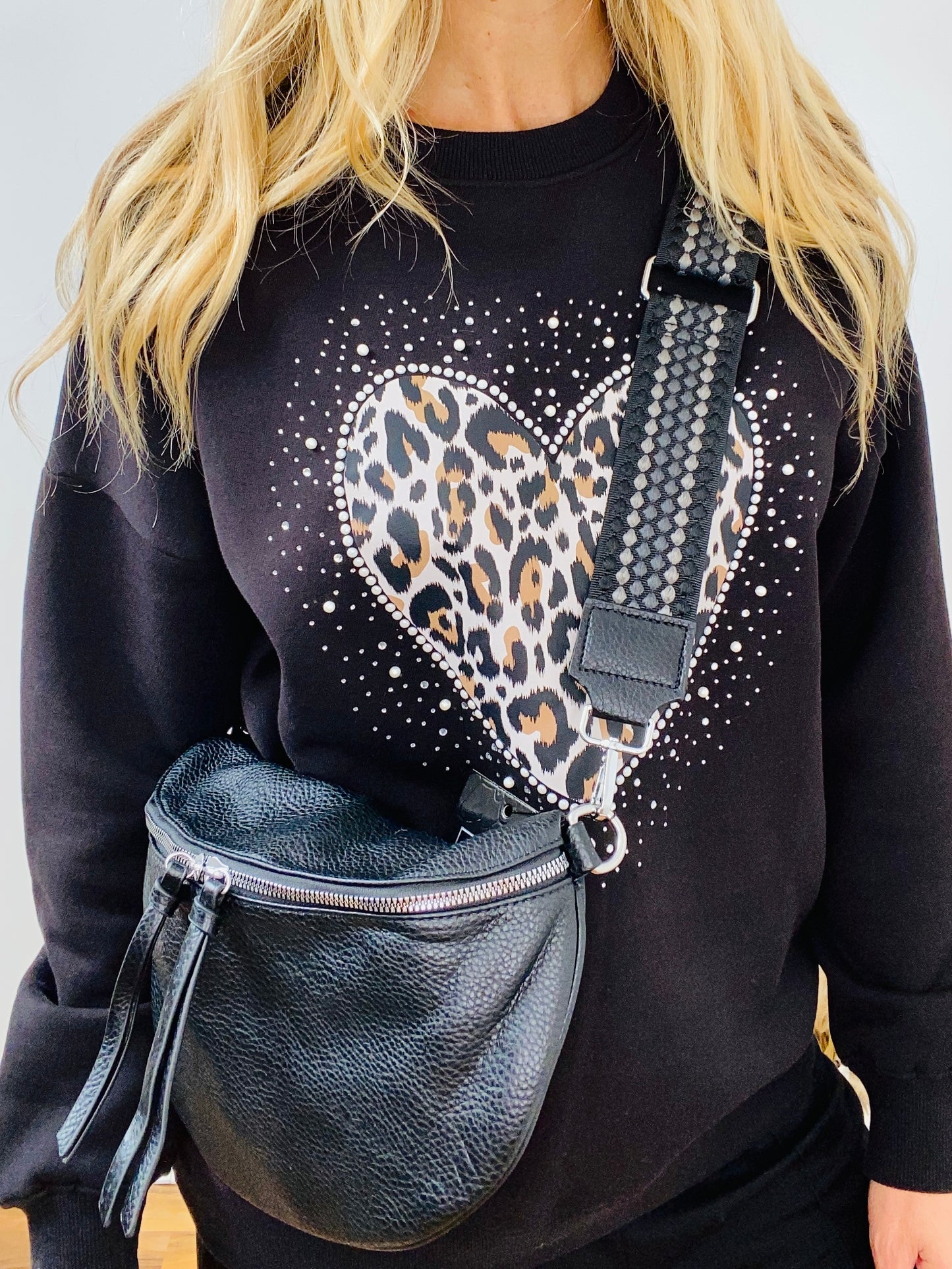 Person wearing a black sweatshirt with a heart design, holding a faux leather black crossbody bum bag