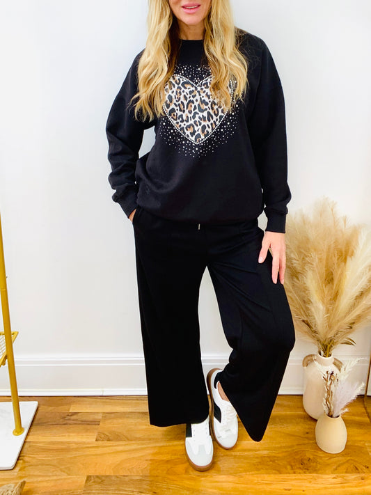 Woman wearing a black sweatshirt with leopard love heart and black joggers and trainers.